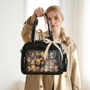 Peut inclure: Un sac à main en cuir verni noir doté d'une poche frontale transparente remplie de divers petits porte-clés représentant des personnages de style anime. Le sac comprend deux anses, une poche latérale et un nœud en ruban crème sur une anse.
