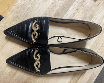 May include: A pair of black leather flats with pointed toes. The vamp features a faux crocodile texture and a gold-colored chain detail. The shoes have a slip-on design and a neutral-colored insole.