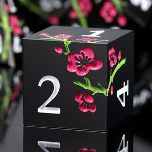 May include: A black die with white numbers and pink flower accents. The die features a floral design with raised pink blossoms and green stems. The number "2" is prominently displayed on one side.
