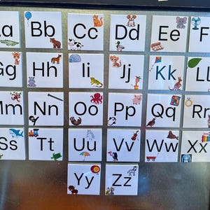 May include: A set of white square alphabet tiles arranged on a metallic surface. Each tile features an uppercase and lowercase letter paired with a colorful illustration representing that letter, such as an apple for Aa, a bear for Bb, and a zebra for Zz.
