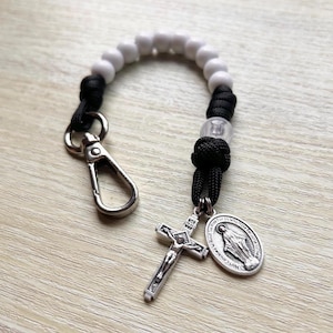 May include: A small rosary keychain with white beads, black cord, and a silver crucifix and oval pendant. The pendant shows the Virgin Mary. A silver clasp is attached for easy use.