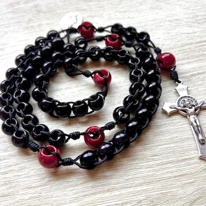 May include: A black rosary with red beads and a silver crucifix. The rosary features black beads connected by knotted black string, with larger red beads interspersed. The silver crucifix has a detailed depiction of Jesus and the letters INRI.