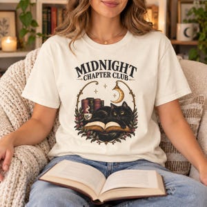 Könnte beinhalten: Ein cremefarbenes T-Shirt mit einer Grafik einer schwarzen Katze, die auf einem aufgeschlagenen Buch liegt. Das Design enthält den Text "MIDNIGHT CHAPTER CLUB", eine Mondsichel, eine dampfende Tasse und einen kreisförmigen Blumenrahmen mit Büchern.