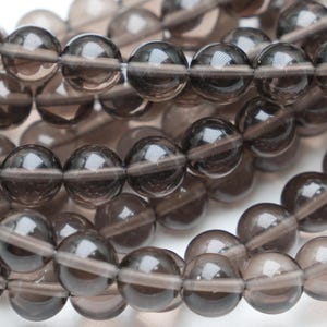 Peut inclure: Gros plan sur des brins de perles de pierres précieuses en quartz fumé. Les perles sont rondes, translucides et de couleur gris-brun foncé. Les perles sont enfilées sur un cordon fin et transparent, adaptées à la création de bijoux.