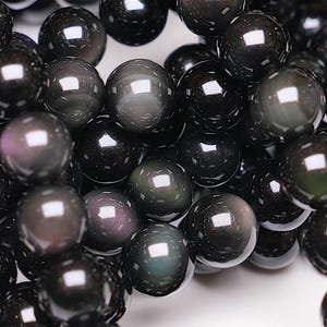 Può includere: Primo piano di una collana di perle di ossidiana nera. Le perle sono rotonde e hanno una finitura liscia e lucida. L'ossidiana è una pietra scura e opaca con un leggero bagliore iridescente.