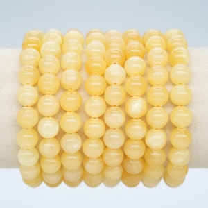 Peut inclure: Plusieurs bracelets composés de perles rondes jaune clair. Les perles sont lisses et polies, enfilées sur un élastique. Les bracelets sont présentés sur une surface blanche sur un fond bleu clair. Les perles mesurent environ 0,8 cm de diamètre.