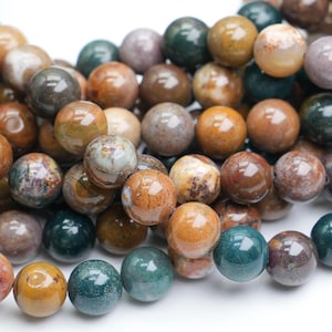 Puede incluir: Primer plano de una colección de cuentas redondas de piedras preciosas en varios colores. Las cuentas varían del verde azulado oscuro y verde bosque a tonos de marrón, beige y malva. Las cuentas están ensartadas, para la fabricación de joyas.
