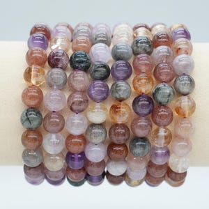 Può includere: Una collezione di braccialetti di perline con una varietà di colori, tra cui viola, arancione, marrone e grigio. Le perline rotonde sembrano essere fatte di un materiale traslucido. I braccialetti sono esposti su una superficie color crema.
