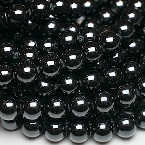Puede incluir: Primer plano de una hebra de cuentas de hematites negras. Las cuentas son redondas y lisas, con un alto pulido. Están dispuestas en un patrón aleatorio, creando una textura visualmente atractiva.