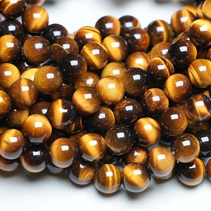 Può includere: Una collana di perle rotonde in occhio di tigre lucidate in tonalità di marrone e oro. Le perle hanno una lucentezza naturale e iridescente.