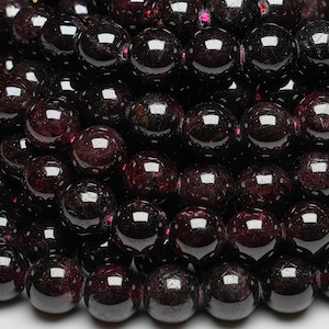 Può includere: Primo piano di una collana di perle di pietre preziose rosso scuro. Le perle sono rotonde e lisce, con un'alta lucidatura. Sono disposte in uno schema casuale, creando una texture visivamente accattivante.