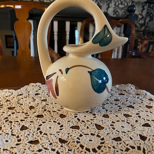 May include: A cream-colored ceramic pitcher with a curved handle and a spout. The pitcher is decorated with blue and green leaf designs and a maroon floral accent. The pitcher is sitting on a cream-colored crocheted doily.