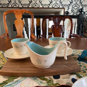 Peut inclure: Une saucière en céramique blanche vintage et des tasses à thé assorties avec un intérieur bleu clair et un motif floral. La saucière est sur une planche de service en bois, avec les tasses à thé sur des soucoupes.