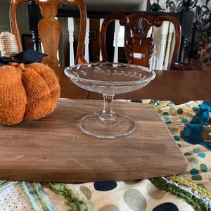May include: A clear glass dessert dish with a decorative leaf pattern around the rim. The dish sits on a wooden cutting board, with a decorative pumpkin and patterned fabric in the background. The dish is approximately 5 inches in diameter.