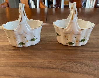May include: Two white porcelain baskets with handles, decorated with a daisy pattern and gold trim. The baskets are on a wooden surface. The handles are arched and have a gold accent.
