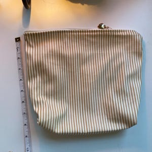May include: A beige and white striped zippered pouch. The pouch is approximately 11 inches long. The zipper pull is gold-colored. The pouch is made of a fabric with vertical stripes. The pouch is on a white surface.