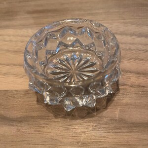 Op de afbeelding: Een heldere, geslepen glazen kom met een stervormig patroon op de bodem en ruitvormige facetten aan de zijkanten. De kom is cirkelvormig en staat op een houten oppervlak.