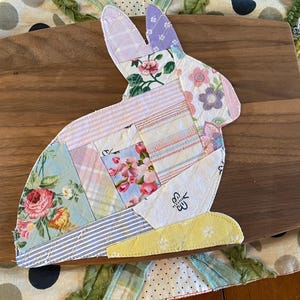 May include: A patchwork bunny-shaped trivet with a variety of floral and striped fabric patterns. The bunny is set on a wooden cutting board, with a patterned fabric border in the background. The trivet is a mix of pastel colors.
