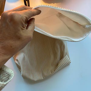 May include: Open, striped pouch with a white zipper. The pouch features vertical tan and white stripes on the exterior and a white interior. The pouch is open, revealing the interior lining.