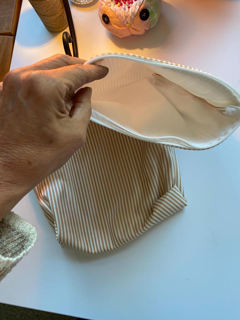 May include: Open, striped pouch with a white zipper. The pouch features vertical tan and white stripes on the exterior and a white interior. The pouch is open, revealing the interior lining.