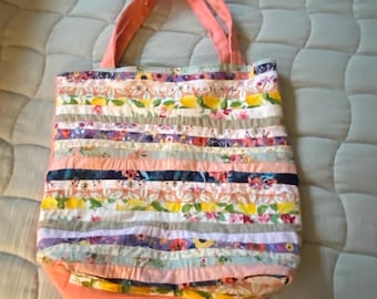 May include: A patchwork tote bag with a floral design. The bag is made from horizontal strips of fabric in various colours and patterns, including pink, yellow, and purple. It has a peach-coloured base and matching handles.