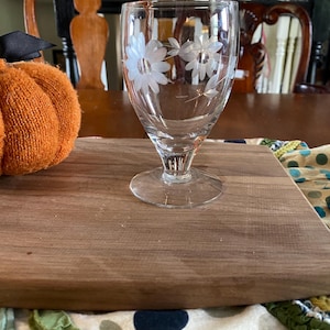 May include: A clear glass goblet with a floral design etched on the bowl. The goblet sits on a round base and has a stem. The glass is on a wooden cutting board with a decorative pumpkin and a patterned cloth.