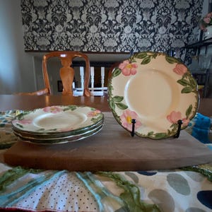 Può includere: Un set di piatti floreali vintage con fiori rosa e foglie verdi. I piatti sono esposti su una tavola di legno, con una tovaglia fantasia in primo piano. I piatti sono supportati da supporti in metallo nero.