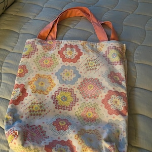 Könnte beinhalten: Eine Stoff-Tragetasche mit einem Blumenmuster in Rosa, Gelb, Blau und Grün. Die Tasche hat einen hellrosa Griff. Die Tasche ist aus einem weichen, gesteppten Material gefertigt.