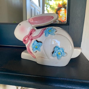 Puede incluir: Figura de conejo de cerámica blanca con detalles rosas en las orejas y un lazo rosa. El conejo tiene recortes de flores azules y está sobre una superficie azul oscuro. Un artículo decorativo de Pascua.