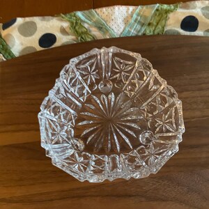 May include: A clear, triangular-shaped glass dish with a detailed starburst and geometric pattern. The dish is sitting on a wooden surface. The dish is likely used for serving small items.