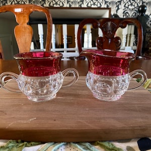 以下が含まれることがあります： ルビーレッドの上部とクリアなエンボス加工が施された下部が特徴の、ヴィンテージガラス製のクリーマーとシュガーボウルのセット。各ピースにはハンドルが付いています。木製のまな板の上に置かれています。