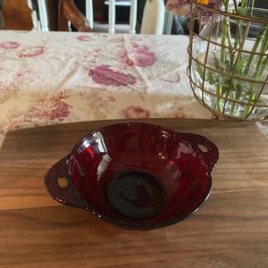 May include: A ruby red glass bowl with two handles, set on a wooden surface. The bowl has a ribbed design and a rounded shape. The background includes a floral tablecloth and a vase of flowers.