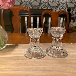 May include: Two clear glass candle holders with a ribbed design. The candle holders are on a wooden surface. A vase with pink flowers is visible in the background.