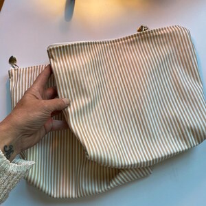 May include: Two beige and white striped zippered pouches. The pouches are rectangular with a rounded bottom. The zipper pulls are silver. The pouches are made of a soft fabric.