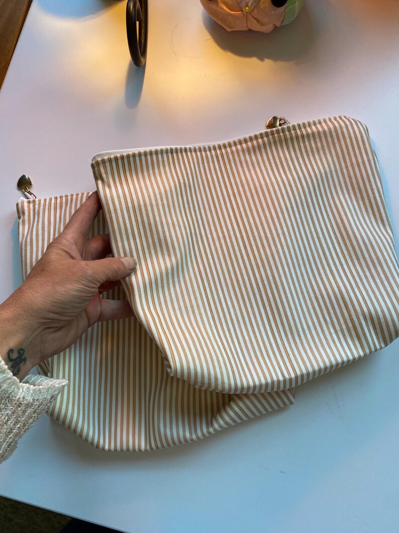 May include: Two beige and white striped zippered pouches. The pouches are rectangular with a rounded bottom. The zipper pulls are silver. The pouches are made of a soft fabric.