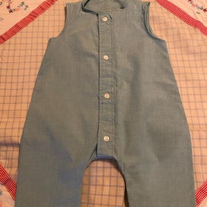 May include: A light blue, sleeveless romper with a button-up front. The romper has a round collar and short legs. The buttons are white. The romper is made of a light fabric.