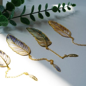 Könnte beinhalten: Vier federförmige Lesezeichen mit goldfarbenen Verzierungen und Ketten. Die Federn haben einen Farbverlauf, darunter Lila, Grün und Braun. Jedes Lesezeichen hat einen kleinen Federanhänger am Ende der Kette.