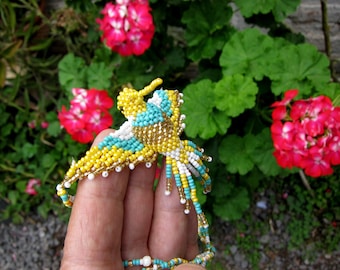 May include: A beaded hummingbird necklace with yellow, white, turquoise, and gold beads. The necklace is being held by a hand against a background of green leaves and pink flowers.