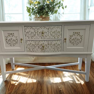 May include: A white painted buffet with two drawers and two cabinet doors. The drawers and doors feature a decorative stencil design in a contrasting color. The buffet has brass-colored hardware and is supported by angled legs with an X-shaped support.