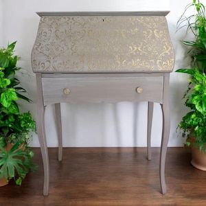 May include: A gray secretary desk featuring a drop-front lid with a gold damask pattern. The desk has a single horizontal drawer with two small, round, gold-toned floral knobs and stands on four slender, curved legs with a scalloped apron.
