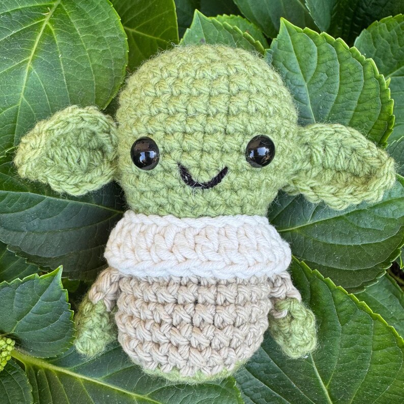 May include: A crocheted green doll with large ears, black eyes, and a smiling mouth. The doll wears a cream-colored collar and a tan outfit. The doll is set against a backdrop of green leaves.