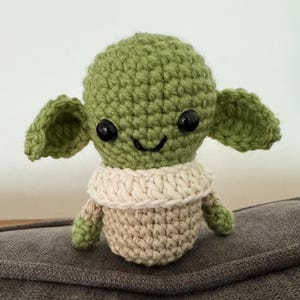 May include: A handmade crochet doll in the likeness of a popular character. The doll features a green head with large ears, black eyes, and a small smile. It has a tan body and a cream-colored collar.