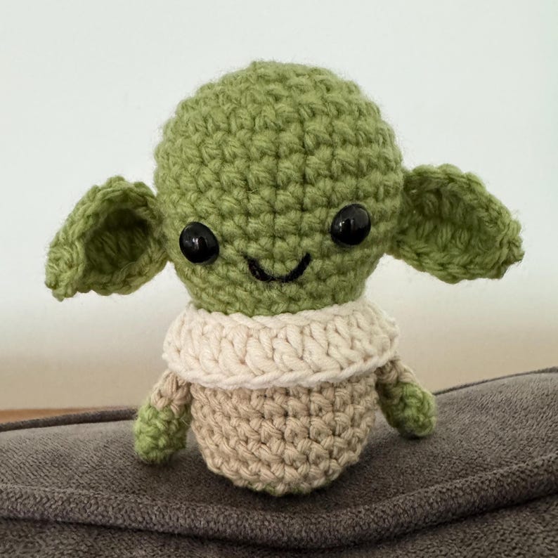 May include: A handmade crochet doll in the likeness of a popular character. The doll features a green head with large ears, black eyes, and a small smile. It has a tan body and a cream-colored collar.