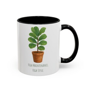 Könnte beinhalten: Eine weiße Keramiktasse mit schwarzem Rand und Henkel. Die Tasse zeigt eine grüne Topfpflanze mit dem Text "High Maintenance. High Style."