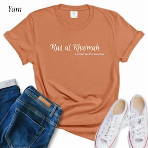 May include: A rust-colored t-shirt with the text "Ras al Khaimah, United Arab Emirates" in a white script. The shirt is paired with blue jeans and white canvas sneakers, creating a casual outfit.