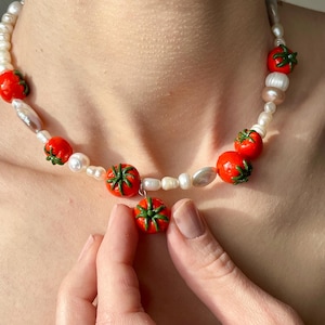 Pode incluir: Um colar feito à mão com contas vermelhas em forma de tomate com detalhes verdes, intercaladas com pérolas brancas e contas de formato irregular. O colar é enfiado em um cordão branco e é exibido em um fundo de tom de pele.