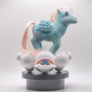 Könnte beinhalten: Ein hellblaues Spielzeugpferd mit rosa Haaren und Flügeln, das auf einer wolkenförmigen Basis steht. Die Basis hat einen Regenbogen und die Worte "Wind Whistler". Das Pony hat rosa und blaue Markierungen an der Flanke.