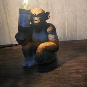 Puede incluir: Lámpara con forma de mono marrón que sostiene una bombilla encendida. La lámpara tiene un acabado texturizado y detallado y se encuentra sobre una superficie de madera. La bombilla está encendida, emitiendo un brillo cálido.