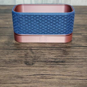 Può includere: Un contenitore decorativo bicolore. La base e l'interno sono color oro rosa, mentre la parte superiore è blu strutturato con un motivo a stella ripetuto. Il contenitore è posto su una superficie di legno.