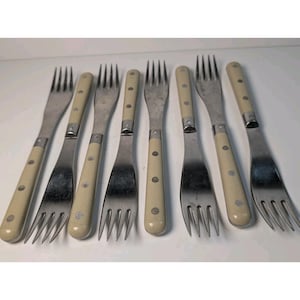 May include: A set of eight vintage forks with stainless steel tines and cream-colored handles. Each fork has three rivets on the handle and a silver metal band where the handle meets the tines. The forks are arranged on a white surface.